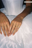 A close-up of a person's hands resting on a white satin dress, wearing a delicate gold band ring with a small clear gemstone