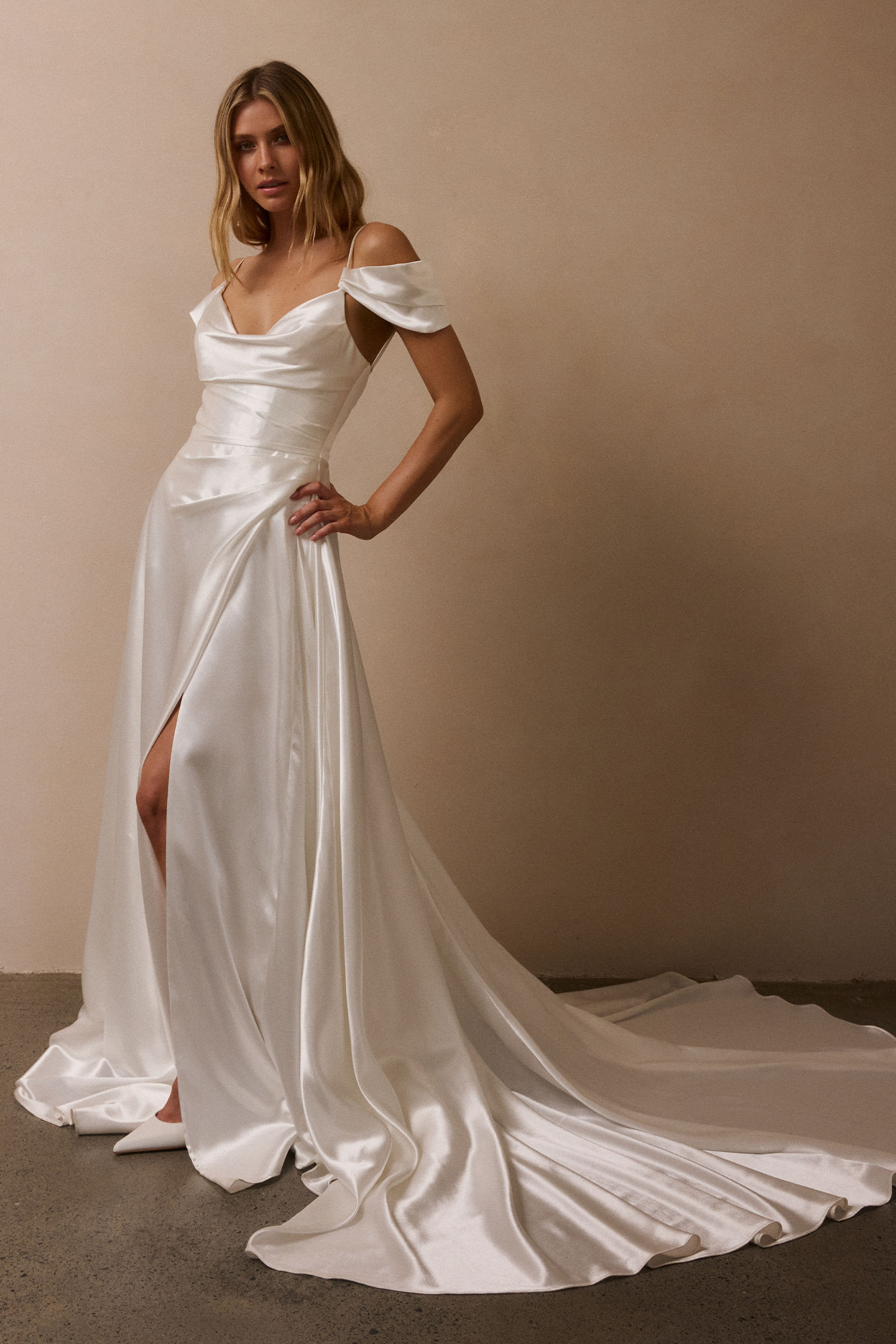 A woman models a white satin off-the-shoulder wedding gown with a thigh-high slit and a long train against a warm beige studio wall_XS_