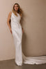 A woman modeling a sleeveless white satin floor-length gown with a high neckline and a long train, standing against a plain beige wall_XS_