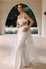 A woman modeling a sleeveless white wedding gown with thin straps, a fitted silhouette, and a long sheer tulle overskirt, standing in a sunlit architectural setting with an archway behind her_XS_