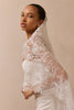 A woman in a white strapless dress wears a sheer white lace bridal veil draped over her shoulders, posed against a solid warm beige studio background.
