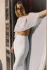 A woman wearing a white two-piece outfit consisting of a sheer flutter-sleeve crop top and a fitted high-waisted midi skirt, posing outdoors next to a shuttered window.