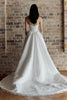 A woman viewed from behind wearing a strapless white wedding gown with a fitted bodice and a full, flowing train against a rustic brick wall.