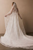 A bride viewed from behind wearing a long white lace wedding dress with a matching sheer lace veil, standing against a plain warm beige wall.