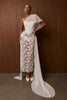 A model wears a dramatic one-shoulder bridal gown featuring a structured white sweetheart bodice, a sheer floral lace skirt, and a long flowing train, posed against a dark wood-paneled wall.