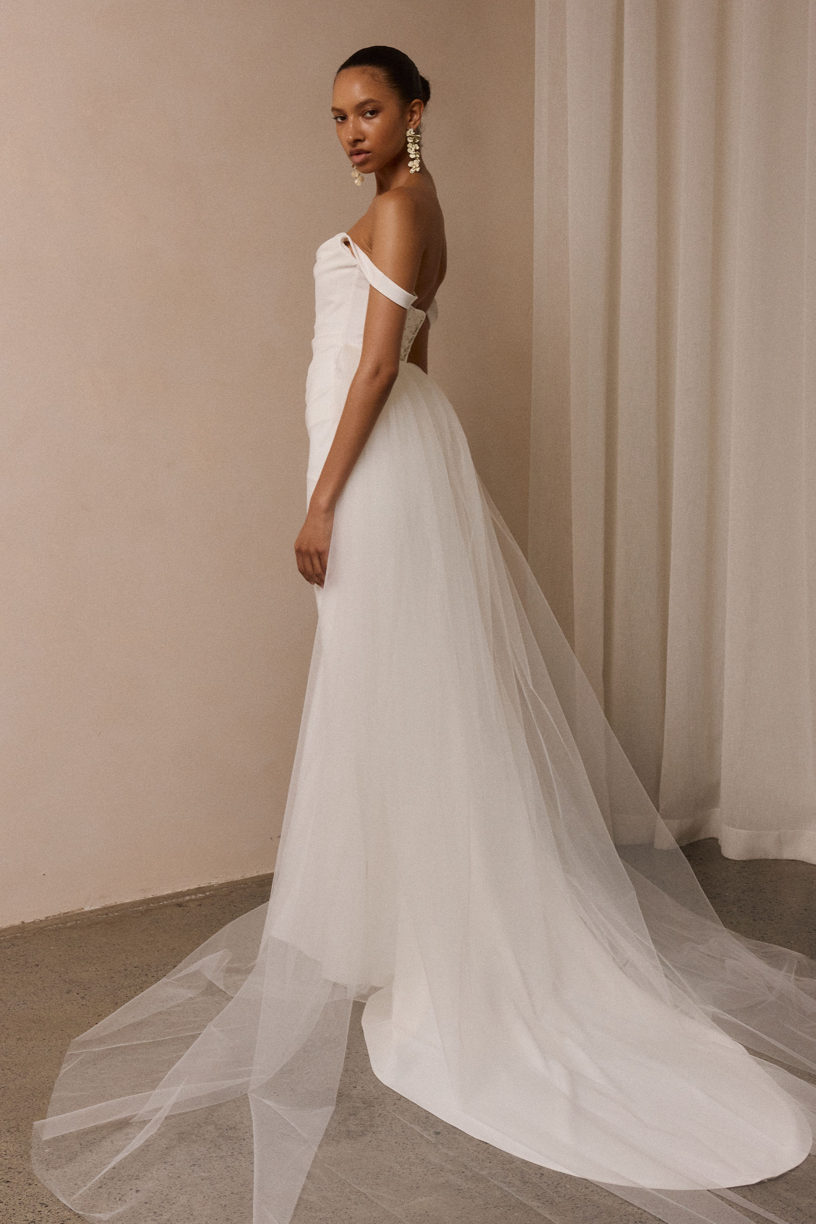 A woman modeling a sleeveless white wedding gown with a fitted bodice, full skirt, and long sheer tulle train against  a plain beige studio wall_XS_