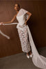 A model wears a white one-shoulder wedding dress featuring a textured corset bodice, a sheer floral lace skirt, and a long flowing train, posed against a dark wood-paneled wall.