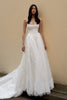 A woman modeling a strapless white wedding gown with a fitted bodice, full lace skirt, and long train against a plain beige wall.