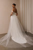 A woman wearing a bridal gown stands with her back to the camera in an indoor setting with beige walls and draped curtains. The dress has an off-the-shoulder neckline, a fitted bodice, and a voluminous tulle skirt with a long train.