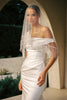 A woman wearing a white off-the-shoulder wedding dress with beaded fringe detailing and a matching sheer veil, standing in an outdoor archway setting.