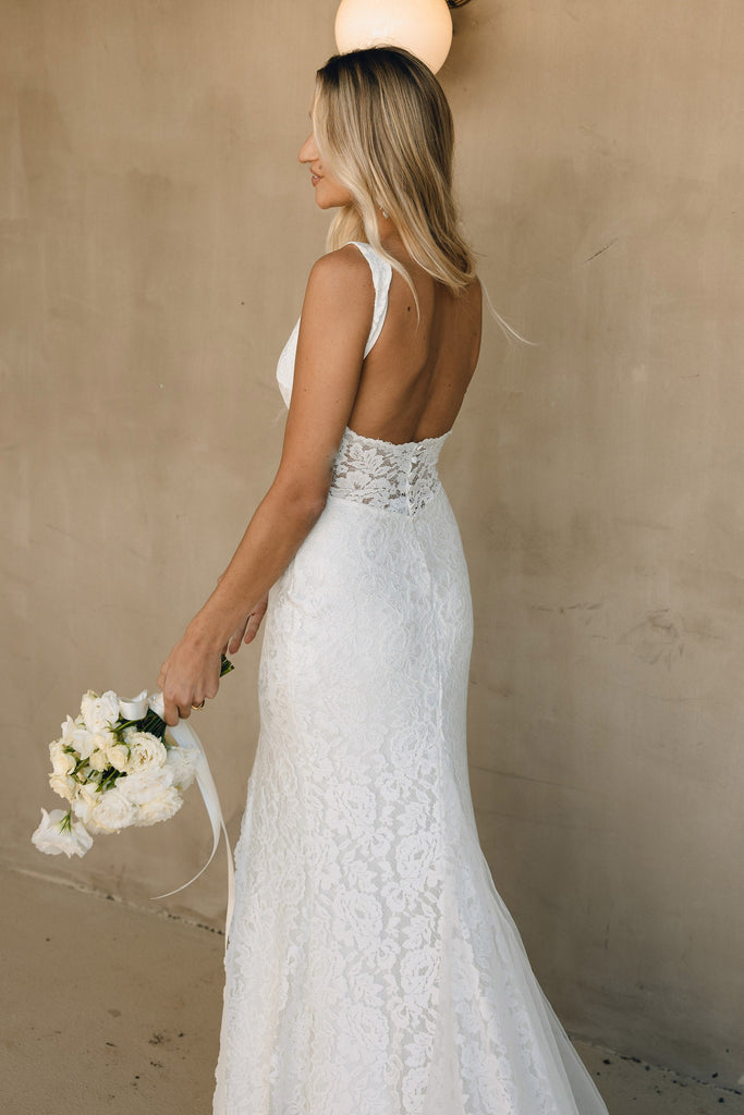 Woman in a lace wedding dress holding a bouquet against a beige wall